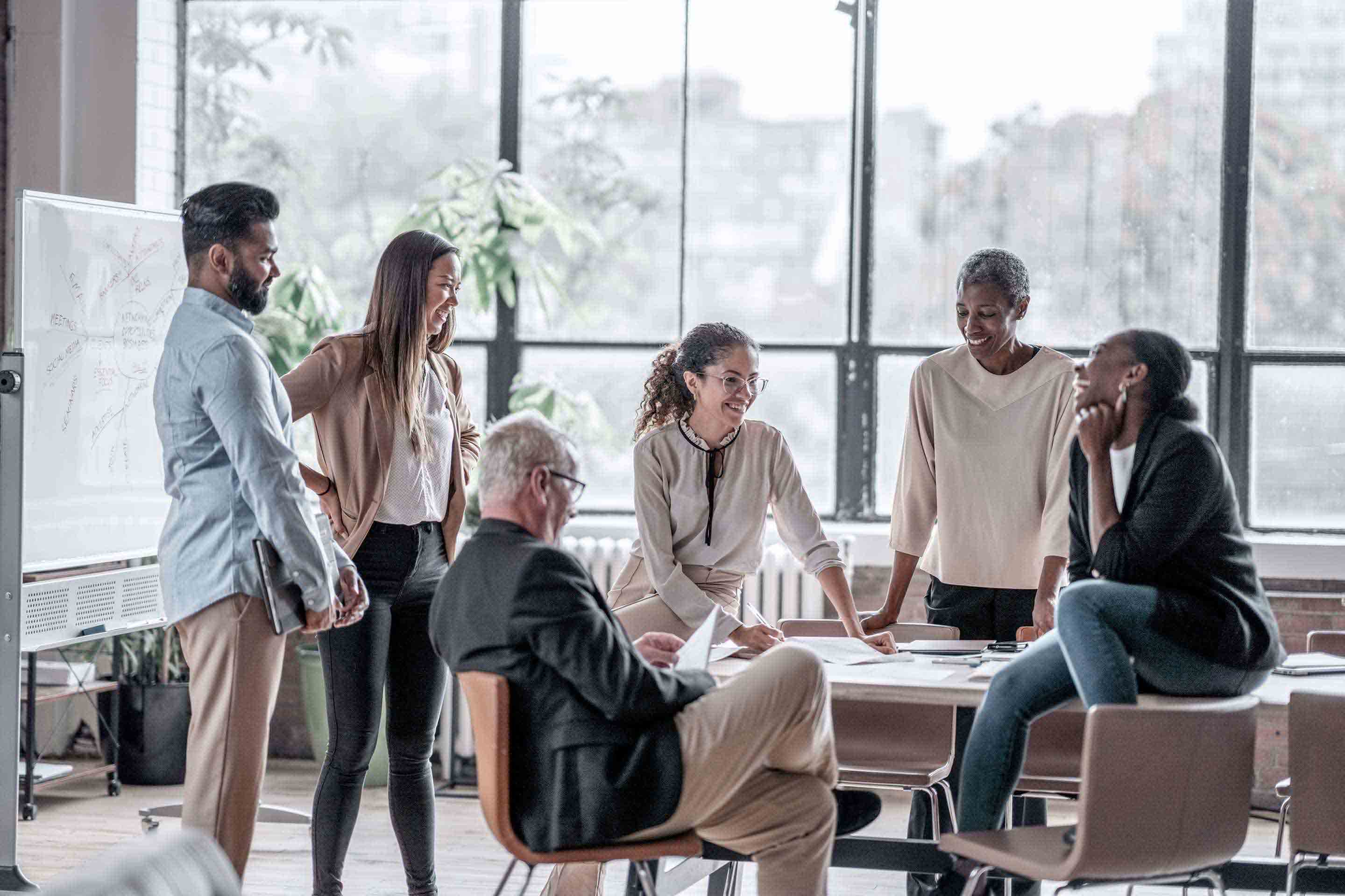 A diverse group of six professionals laugh and engage in a collaborative discussion around a conference table in a modern glass-walled office, with a whiteboard diagram visible in the background. The image represents Honeywell's global workforce and innovative corporate culture.