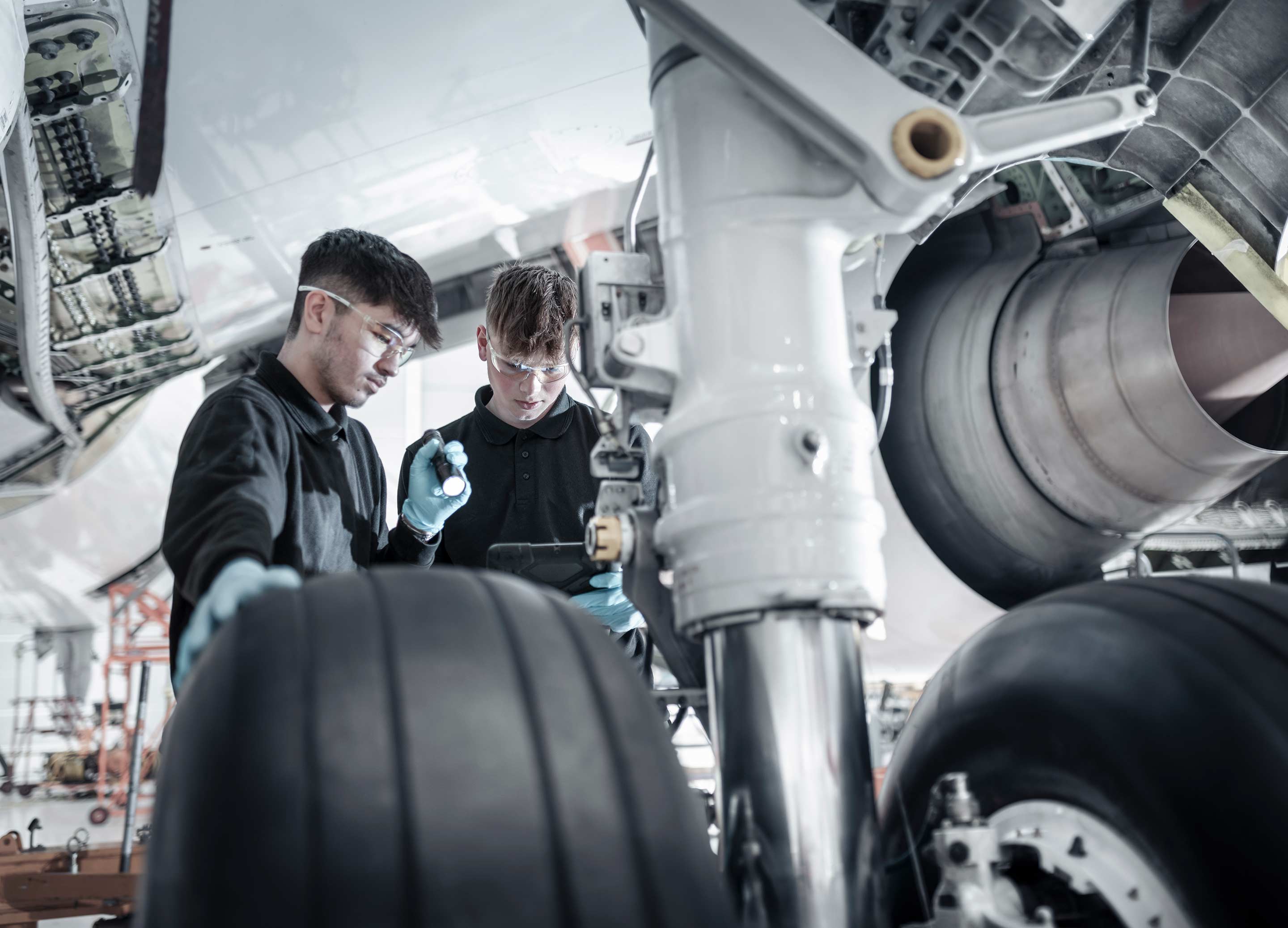 Two aviation maintenance technicians wearing safety glasses and gloves examine the landing gear assembly of a commercial aircraft in a bright MRO hangar, with one using a flashlight. This image reflects Honeywell's landing gear systems, brakes, and wheels product line.