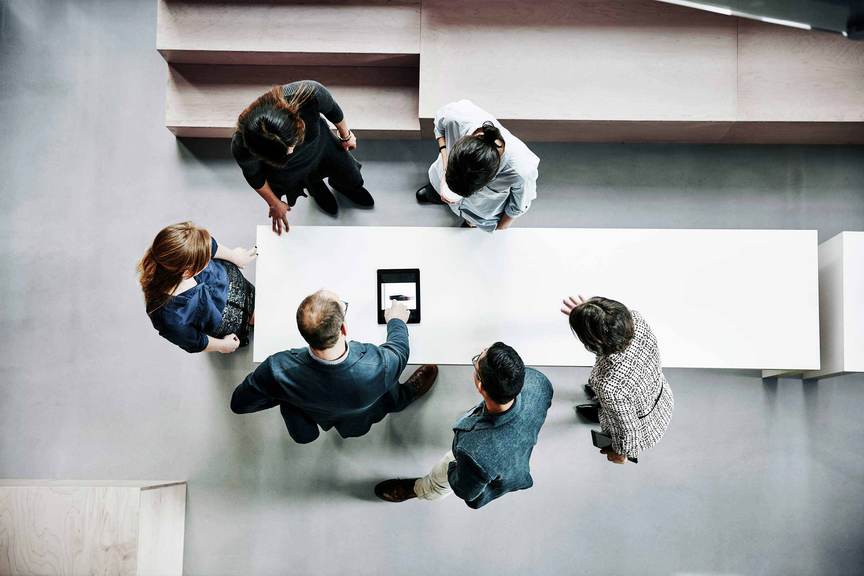 Top-down aerial view of six professionals gathered around a conference table with one person using a tablet, in a modern open-plan office with geometric decor. This image represents Honeywell's global business collaboration and technology integration culture.