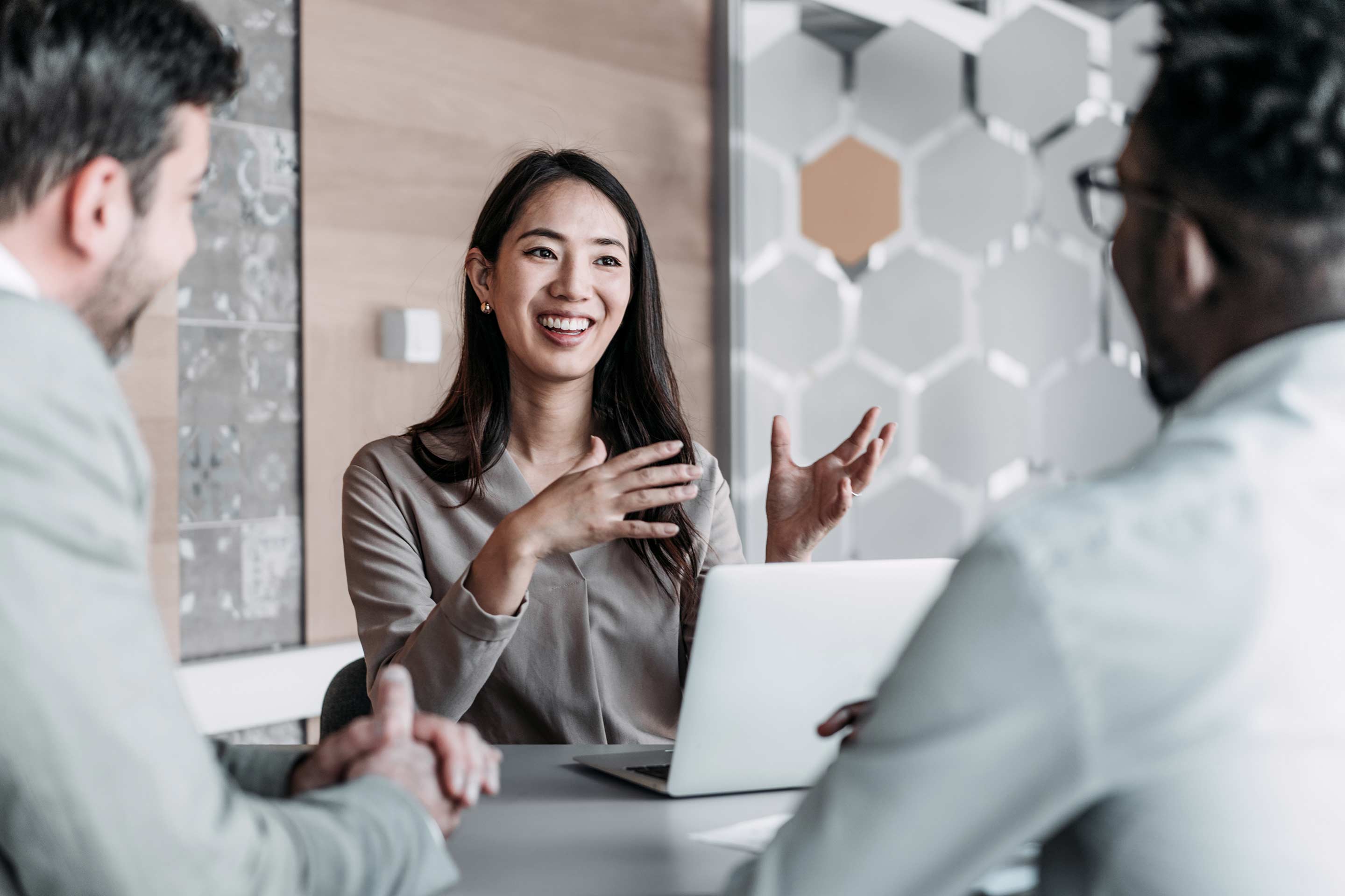 An Asian professional woman smiles and speaks animatedly in a business meeting with two male colleagues around a table with a laptop, in a modern hexagonal-wall office. This image represents Honeywell's inclusive global workforce and customer engagement culture.