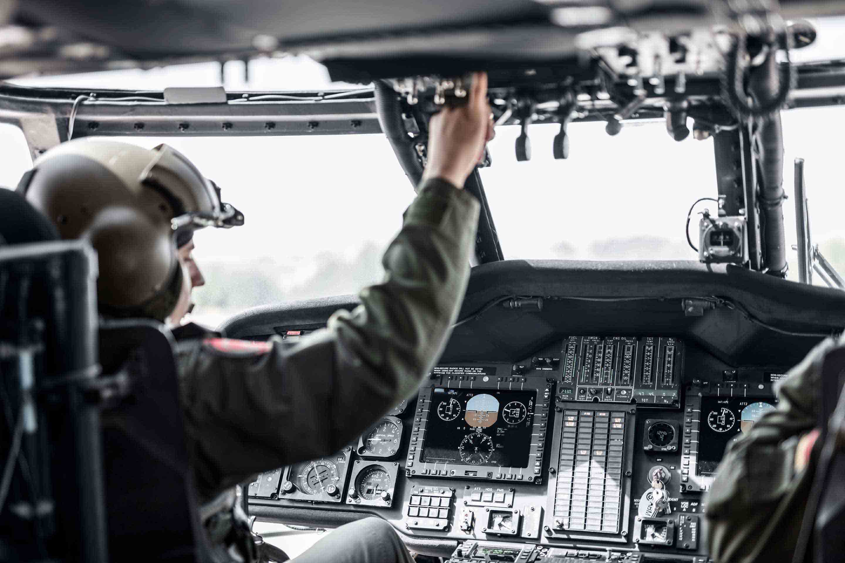Two military helicopter crew members in flight suits and helmets conduct pre-flight checks inside a military helicopter cockpit, with one reaching overhead for switch panel items. This image represents Honeywell's defense avionics, mission systems, and crew alerting technologies.